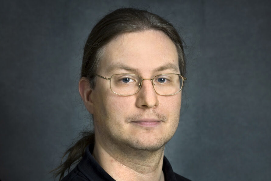 A professional headshot of Paul Hargrove with long brown hair pulled back into a ponytail. He is wearing wire-rimmed glasses and a dark polo shirt against a mottled gray background.