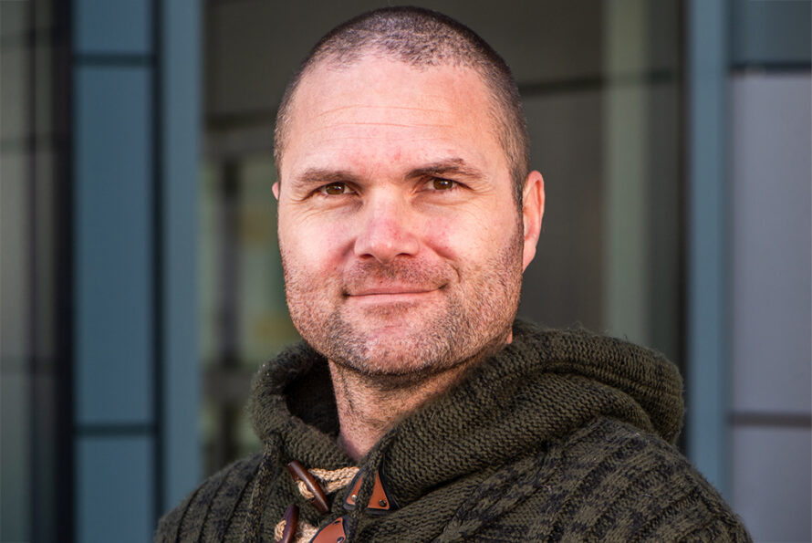 A headshot of Marcus Noack smiling, wearing a dark green knit sweater with wooden toggle buttons, standing against a blurred architectural background.
